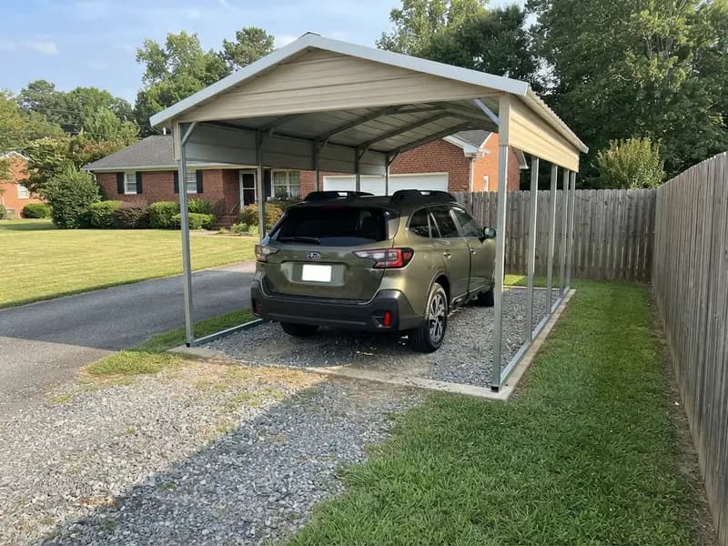 Detached steel carport beside a wood fence sheltering a 2023 Subaru Outback, roof edge clear of the fence