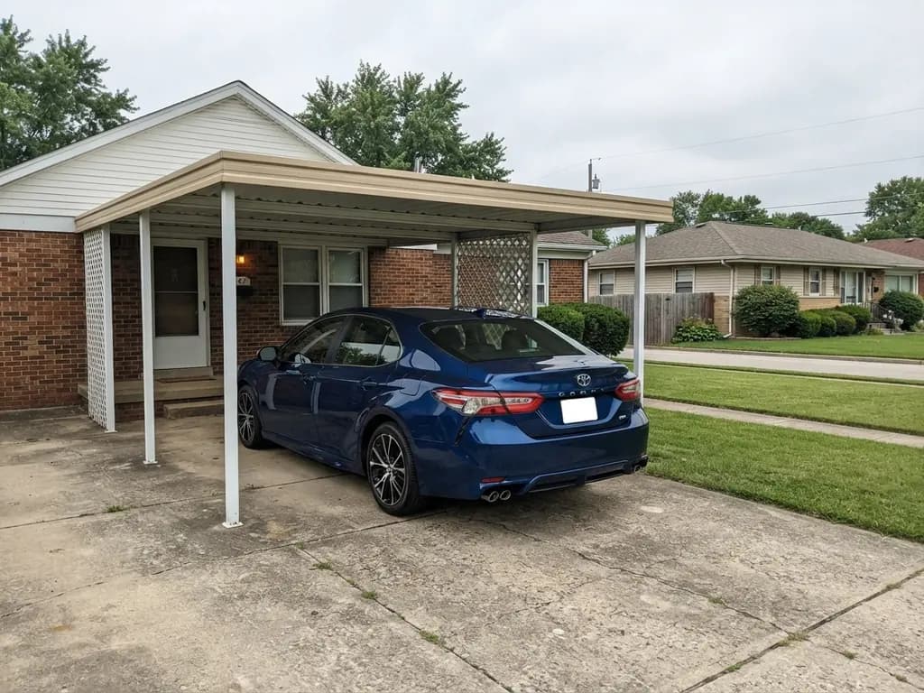 Steel carport open on two sides beside a brick house with a 2023 Toyota Camry parked under its low-slope roof on concrete